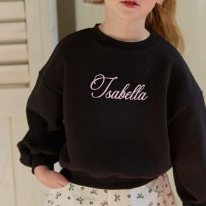 May include: A black long-sleeved sweatshirt featuring a crew neckline and the name Isabella embroidered in a light pink cursive script across the center of the chest. The garment has a relaxed fit with ribbed cuffs and a ribbed hemline.