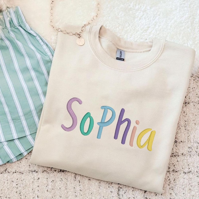 May include: Cream-colored sweatshirt embroidered with the name "Sophia" in colorful thread. The letters are in shades of purple, teal, and yellow. Beside the sweatshirt are striped green shorts and a gold necklace.