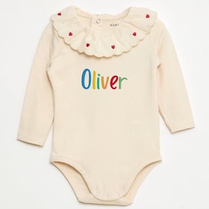 May include: Cream-colored baby bodysuit with long sleeves and a Peter Pan collar. The collar is adorned with small red heart-shaped embroidery. The name "Oliver" is embroidered in colorful letters across the front. A button closure is at the back of the neck.