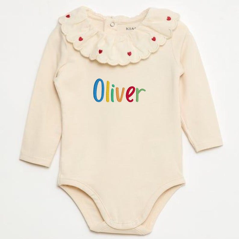 May include: Cream-colored baby bodysuit with long sleeves and a Peter Pan collar. The collar is adorned with small red heart-shaped embroidery. The name "Oliver" is embroidered in colorful letters across the front. A button closure is at the back of the neck.
