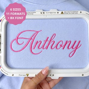 Pode incluir: Um bastidor de bordado de plástico branco emoldurando um tecido azul claro com o nome Anthony bordado em letra cursiva rosa brilhante. Um gráfico circular rosa no canto superior esquerdo exibe o texto 6 SIZES, 11 FORMATS + BX FONT.