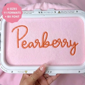 May include: A white embroidery hoop frame containing light pink fabric with the word Pearberry stitched in orange thread. The frame includes text stating 6 SIZES, 11 FORMATS, and BX FONT, along with safety warnings about electronic devices.
