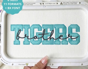 May include: An embroidery design featuring the word TIGERS in a light blue block font with the word brother in a dark gray cursive script layered over it. The design is displayed within a white plastic embroidery hoop on ribbed fabric.