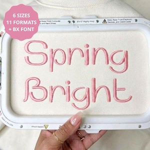 Peut inclure: Un tambour à broder contenant un tissu blanc avec le texte brodé en rose Spring Bright. Le cadre du tambour porte les mentions 6 SIZES, 11 FORMATS + BX FONT, 8x13 Mighty Hoop, et Keep Away From Electronic Devices, and Credit Cards.