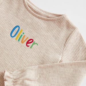 May include: Beige ribbed long-sleeve shirt with the name "Oliver" embroidered in rainbow colors across the chest. The shirt has a round neckline and a ruffled cuff detail on the sleeve.