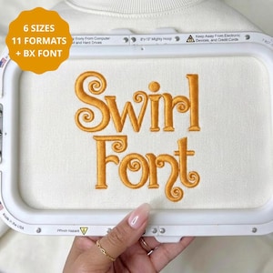 May include: An embroidery hoop frame containing white fabric with the words Swirl Font stitched in gold thread. An orange circular graphic in the top left corner displays the text 6 SIZES 11 FORMATS + BX FONT. The frame is an 8x13 inch Mighty Hoop.