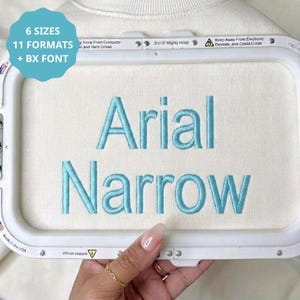 Pode incluir: Uma peça de tecido branco presa em um bastidor de bordado branco, com o texto bordado em azul claro Arial Narrow. Um gráfico circular azul-petróleo no canto superior esquerdo exibe o texto 6 SIZES 11 FORMATS + BX FONT em branco.