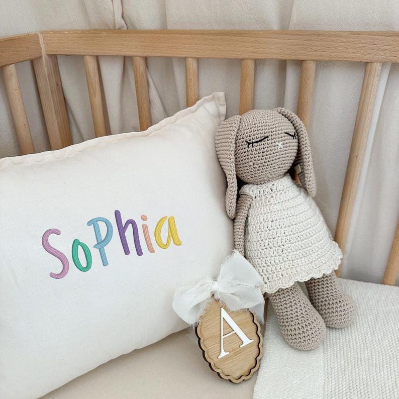 May include: A crocheted bunny doll in a white dress and a pillow embroidered with the name "Sophia" in pastel colors. A wooden tag with the letter "A" is attached with a white ribbon. The items are on a crib.