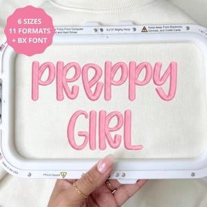 Könnte beinhalten: Ein Stickrahmen mit weißem Stoff und dem rosa gestickten Text PREPPY GIRL. Eine rosa kreisförmige Grafik in der oberen linken Ecke zeigt den Text 6 SIZES 11 FORMATS + BX FONT. Der Rahmen enthält aufgedruckte Sicherheitshinweise.