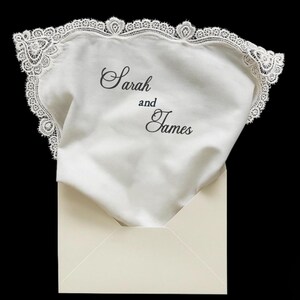May include: A white fabric handkerchief with a decorative lace border, featuring the black printed text Sarah and James. The handkerchief is partially tucked into a cream-colored envelope against a solid black background.
