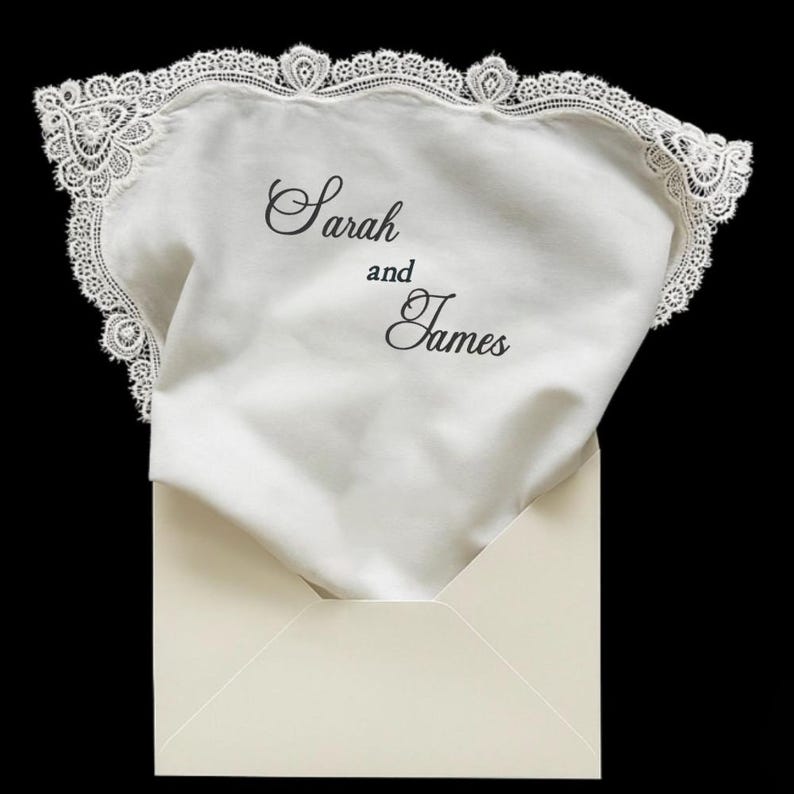 May include: A white fabric handkerchief with a decorative lace border, featuring the black printed text Sarah and James. The handkerchief is partially tucked into a cream-colored envelope against a solid black background.