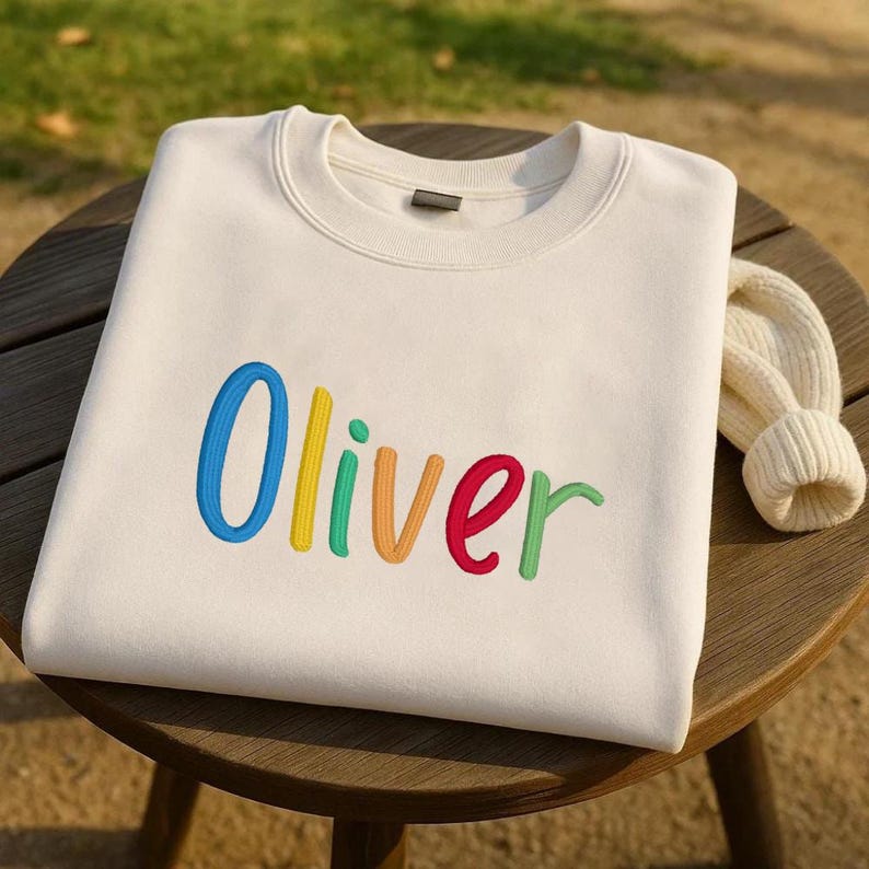 May include: A cream-colored sweatshirt with the name "Oliver" embroidered in colorful letters. The letters are blue, yellow, green, orange, and red. A folded cream-colored knit hat rests on the wooden surface.