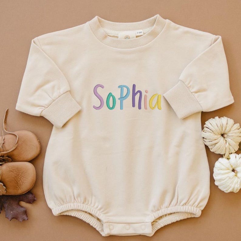 May include: Cream-colored baby romper with long sleeves and a round neckline. The name "Sophia" is embroidered across the chest in colorful thread. The romper has ribbed cuffs and a snap closure at the bottom. A pair of baby shoes and decorative pumpkins are in the image.