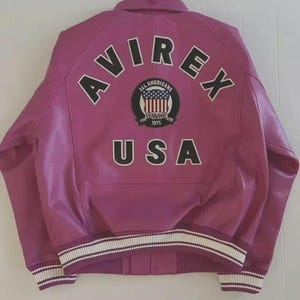 May include: A magenta leather jacket with the word "AVIREX" arched across the shoulders in black block letters. Below the word "AVIREX" is a circular patch with the words "ALL AMERICANS" and "ESTABLISHED 1975". The word "USA" is below the patch.