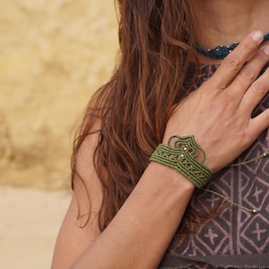 May include: A green macrame bracelet with a decorative design and gold-colored beads. The bracelet is worn on a wrist. A matching green macrame necklace is also visible. The person is wearing a brown top.
