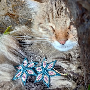 Puede incluir: Un par de pendientes de flores de macramé turquesa y lavanda con ganchos plateados. Los pendientes se colocan sobre un gato atigrado durmiendo con los ojos cerrados, descansando al sol.