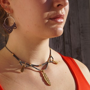 Peut inclure: Une femme porte un ensemble de bijoux faits à la main. L'ensemble comprend un collier en macramé gris foncé avec des accents dorés et un pendentif en forme de plume, ainsi que des boucles d'oreilles créoles assorties. La femme porte un haut rouge.