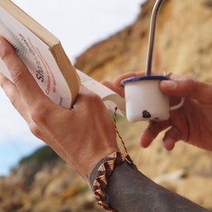 Puede incluir: Una persona sostiene un libro y una pequeña taza de esmalte blanco con una pajita de metal. La taza tiene un borde azul y un diseño negro. Se ve una pulsera tejida con patrones marrones y beige.