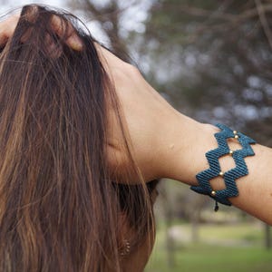 Puede incluir: Una pulsera de macramé turquesa con diseño geométrico y cuentas doradas. La pulsera se luce en una muñeca, con pelo largo castaño al fondo. Un accesorio hecho a mano.
