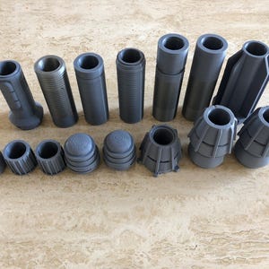 May include: A selection of grey 3D-printed components, including cylindrical tubes, threaded rods, and cone-shaped pieces. The parts vary in size and design, with some featuring ridges and others smooth surfaces. The objects are arranged on a light-coloured surface.