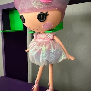 May include: A Lalaloopsy doll with a pink and white dress, pink shoes, and a pink bow. The doll has button eyes and rosy cheeks. The doll is standing in front of a purple and green shelf.