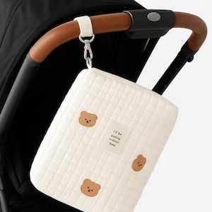 以下が含まれることがあります： クリーム色のベビーカーオーガナイザーバッグ。茶色のクマのアップリケが3つと「I'll be putting individuals best」の文字が入っています。茶色の革のハンドルが付いたベビーカーに取り付けられています。