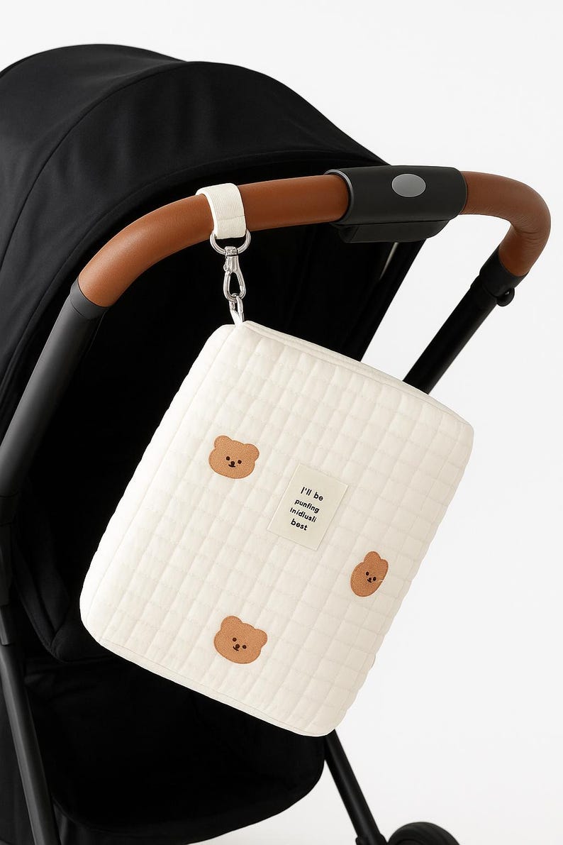 以下が含まれることがあります： クリーム色のベビーカーオーガナイザーバッグ。茶色のクマのアップリケが3つと「I'll be putting individuals best」の文字が入っています。茶色の革のハンドルが付いたベビーカーに取り付けられています。