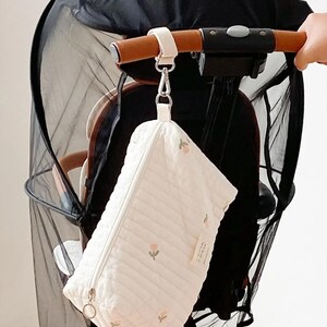 以下が含まれることがあります： 白のキルティングベビーカーオーガナイザーバッグ。ジッパー開閉と小さなピンクの花柄刺繍が施されています。茶色の革のグリップが付いたベビーカーのハンドルからバッグが吊り下げられています。黒いメッシュキャノピーが見えます。