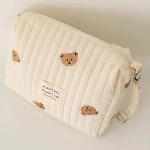 以下が含まれることがあります： クリーム色のキルティング化粧ポーチ。テディベアの刺繍が施されています。ファスナー開閉、テキスト入りの小さな布製ラベル、取り外し可能なストラップ付き。バッグは垂直ステッチでデザインされています。