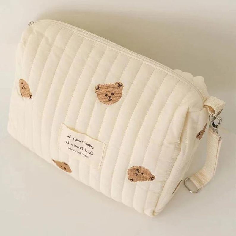 以下が含まれることがあります： クリーム色のキルティング化粧ポーチ。テディベアの刺繍が施されています。ファスナー開閉、テキスト入りの小さな布製ラベル、取り外し可能なストラップ付き。バッグは垂直ステッチでデザインされています。