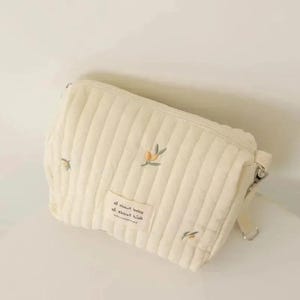 以下が含まれることがあります： クリーム色のキルティングベビーバッグ。オリーブの枝の刺繍が施されています。長方形で、ファスナー開閉と取り外し可能なショルダーストラップが付いています。バッグには「all about baby all about kids」と書かれた小さなラベルが付いています。