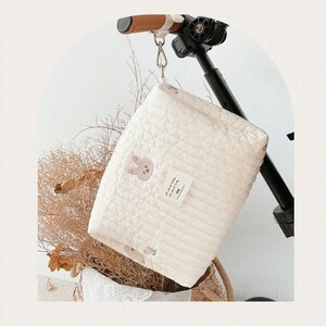 以下が含まれることがあります： キルトデザインのクリーム色のベビーブランケットがベビーカーから吊り下げられています。ブランケットにはウサギの顔の刺繍とテキストの小さなタグが付いています。ベビーカーのハンドルは茶色と黒です。