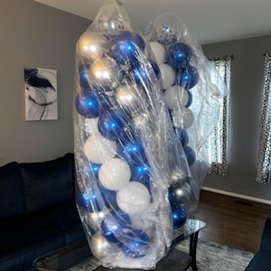 May include: Two large clear plastic bags filled with balloons. The balloons are a mix of white, silver, and blue, some with a metallic sheen. The balloons are likely for a party or event.