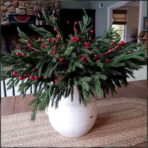 May include: A white ceramic vase filled with evergreen branches and bright red berries. The arrangement sits on a woven jute runner, creating a rustic and festive centerpiece. The vase has a rounded shape and a slightly distressed finish.