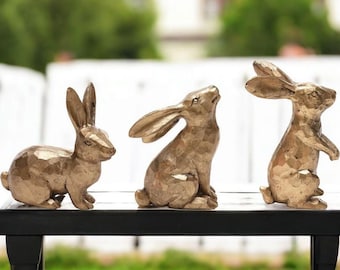 May include: Three bronze-colored rabbit figurines in various poses sit on a black surface. One rabbit is seated, another looks upward, and the third stands with its ears up. The background is blurred, with greenery and a white fence visible.