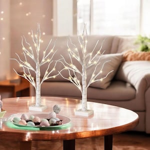 May include: Two decorative white birch trees with warm white LED lights. The trees are on square white bases and are placed on a round wooden table. The table also holds a plate of colorful candies.