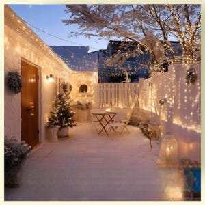 May include: A snow-covered outdoor patio decorated for the holidays. String lights illuminate the white walls, fence, and trees. A small Christmas tree and a table with chairs are visible, creating a cozy, festive atmosphere.