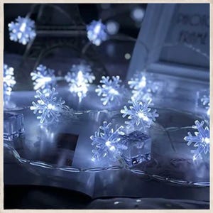 May include: A string of snowflake-shaped LED lights, emitting a cool blue glow. The lights are strung on a thin, silver wire, creating a festive and decorative ambiance. The snowflakes are made of clear plastic and are illuminated with bright white lights.