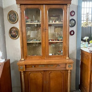 Può includere: Un'antica vetrina in legno con ante in vetro e un mobile inferiore. L'armadio è di colore marrone caldo con accenti dorati sulle colonne e sulle modanature. Le ante in vetro rivelano ripiani pieni di vari piccoli oggetti.