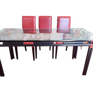 Peut inclure: Une table à manger rectangulaire avec un plateau en verre transparent et un cadre noir doté d'accents rouges aux jointures. Trois chaises rouges à dossier haut sont placées derrière la table, qui possède quatre pieds en doubles tubes noirs.