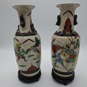 May include: Pair of antique ceramic vases with detailed hand-painted scenes of figures in colorful attire. The vases have a crackle glaze finish, dark brown handles, and sit on dark wooden stands. The overall color palette includes beige, red, green, and blue.