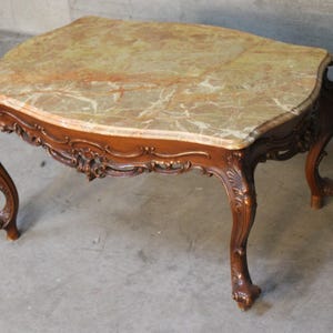 May include: An antique coffee table with a marble top and ornate wooden base. The tabletop features a beige and brown marble pattern. The table has curved legs with detailed carvings, showcasing a classic design. The table is approximately 120 cm long.