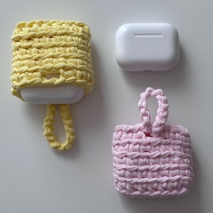 May include: Two crochet AirPod cases, one pale yellow and one pink, sit beside a white AirPod case. The yellow case has a loop, the pink one a small handle. Both are square. The cases are made of a thick yarn.