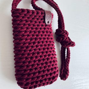 May include: A burgundy-colored, hand-crocheted phone bag with a matching strap and handle. The bag features a textured, woven design and a small, brown leather tag with a logo. The strap is knotted at the end.