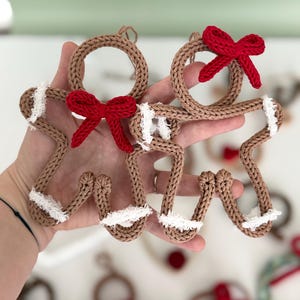 May include: Two gingerbread-shaped ornaments crafted from brown yarn, featuring a circular head, a red yarn bow, and white frayed yarn accents on the arms and legs. Each ornament includes a small loop at the top for hanging purposes.