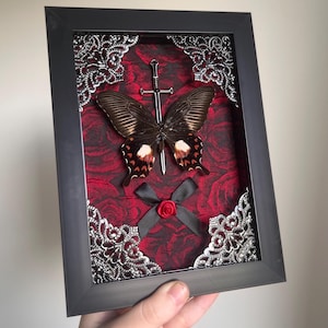 Puede incluir: Obra de arte enmarcada con una mariposa, una espada plateada y un lazo negro con una rosa roja. El fondo es una tela roja oscura con un patrón de rosas, enmarcada por filigranas plateadas ornamentadas.