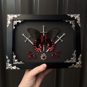 May include: Framed artwork with a black and red butterfly, swords, red gems, and a heart pendant on a black backdrop. The black frame has silver corner details. The artwork is a unique decorative piece.
