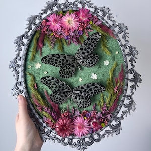 May include: An oval, ornate, silver-colored frame encases a green felt background adorned with two butterflies with black and white patterned wings. The butterflies are surrounded by pink and purple flowers and dried grasses, creating a nature-inspired display.