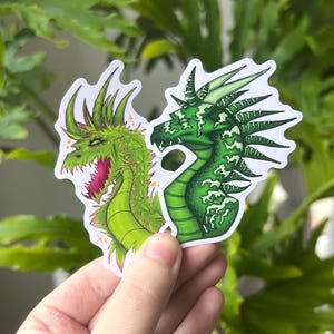 Könnte beinhalten: Zwei Drachenaufkleber. Ein Aufkleber zeigt einen grünen Drachen mit weit geöffnetem Maul und rosa Innenraum. Der andere Aufkleber zeigt einen grünen Drachen mit detailliertem Design. Beide Aufkleber haben einen weißen Rand und werden vor grünen Blättern gehalten.