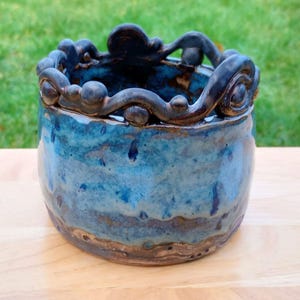 May include: A handcrafted ceramic bowl with a unique, wavy rim design. The bowl features a blend of blue and brown glaze, creating a textured, artistic appearance. The bowl is sitting on a wooden surface.